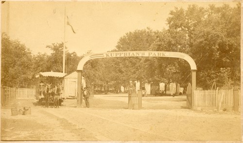A roadway leads under an arch bearing the name Kupfrian's Park. To the left of the arch, a mule-drawn trolley car takes guests away from the park.