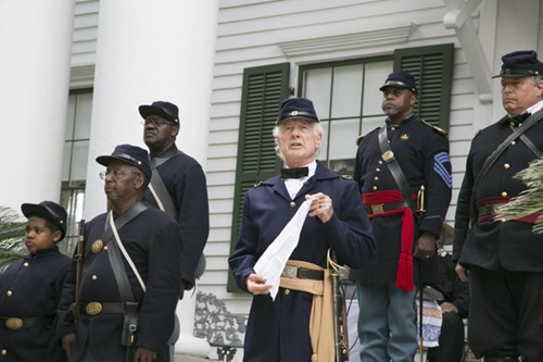 A group of men in recreated Union Army uniforms stand on a porch. The man in the center holds a copy of the Emancipation Proclamation and is reading it aloud. Large white columns of the porch and a window with dark green shutters are seen behind the men.