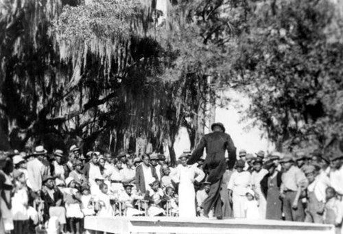 A male performer on a wooden platform captivates a crowd of men, women, and children adorned in white outfits.