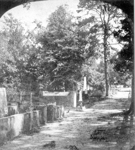 To the left of a wide dirt path are small stone walls. Within the walls are stone burial monuments. A white obelisk is seen in the distance.